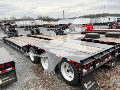 Flatbed trailer showing lowboy deck with multiple axles and tires, chrome wheel rims, and reflective safety tape; background includes other trailers in lot.