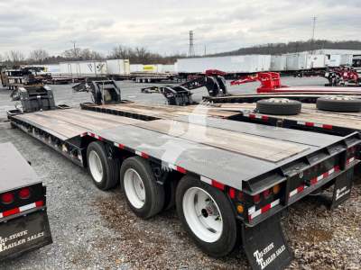 Trailer showing a lowboy flatbed with dual rear axles and metal deck, long trailer in a yard with other trailers and equipment.