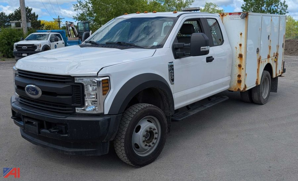 Ford F-450 Super Duty service truck with white cab and rusted utility box on the bed, front grille and bumper visible, side mirrors, running boards, standard steel wheels.