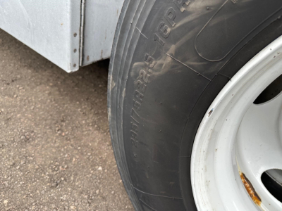 Flatbed trailer wheel and fender area showing tire with visible tread and white rim; portion of metal trailer body in background.