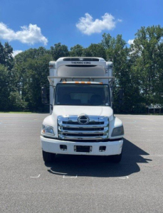 Hino 258/268 reefer truck front view showing Thermo King refrigeration unit on cab roof, chrome grille, white body, clear sky.