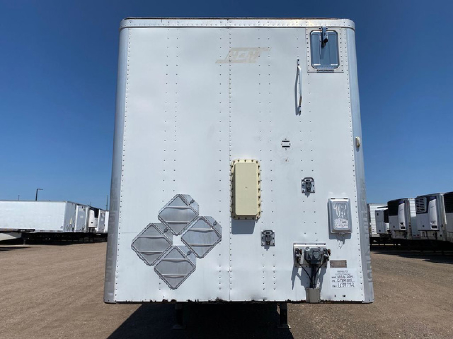 Rear view of a white enclosed utility van trailer showing various decals and access panels. Visible text includes reflective diamond placard, square latch, and small labels.