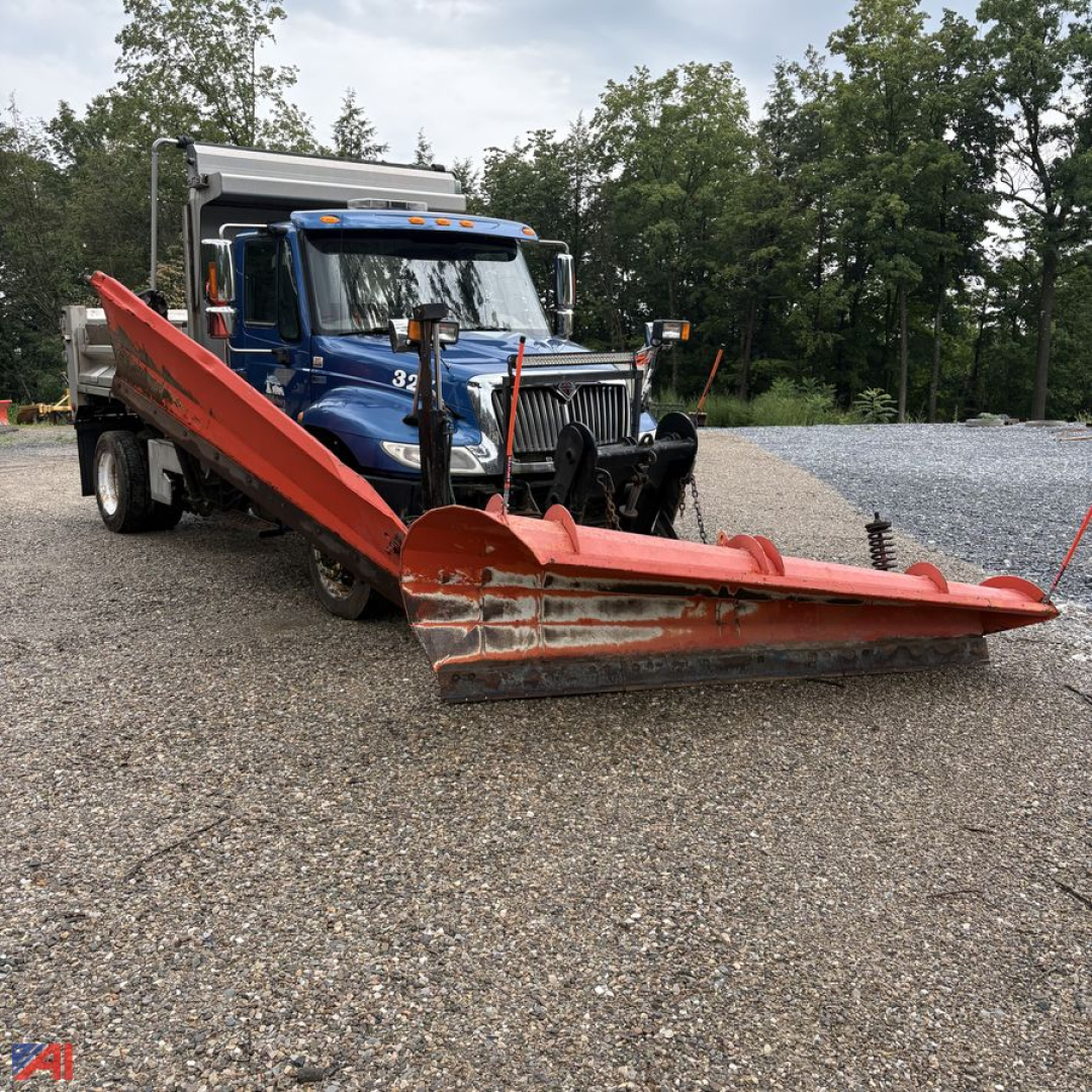 Dump truck with large red snow plow attachment in front, blue International DuraStar 4300, gravel lot, trees in background.
