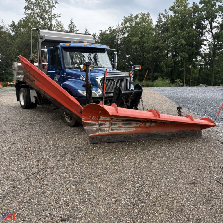 Dump truck with large red snow plow attachment in front, blue International DuraStar 4300, gravel lot, trees in background.