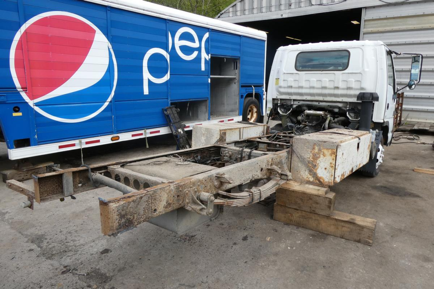 Vehicle chassis with cab and rusted frame, flatbed chassis on ground next to Pepsi trailer in lot.
