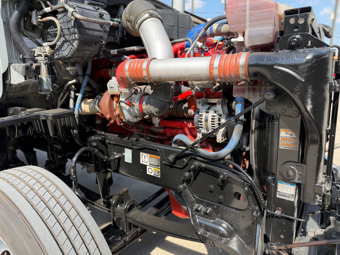 Peterbilt 579 engine bay showing red engine block, intake piping, hoses, coolant reservoir, and wiring in front of the cab
