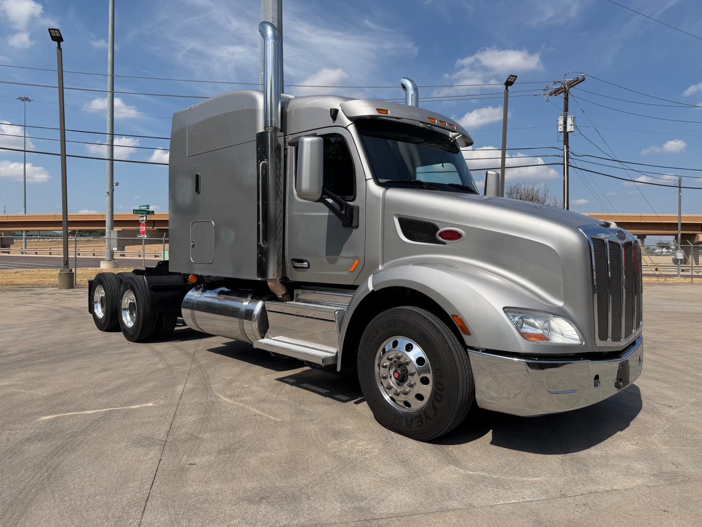 Peterbilt 579 tractor in a parking lot, silver exterior, sleeper cab, chrome exhaust stacks, large front chrome grille, dual rear axles.