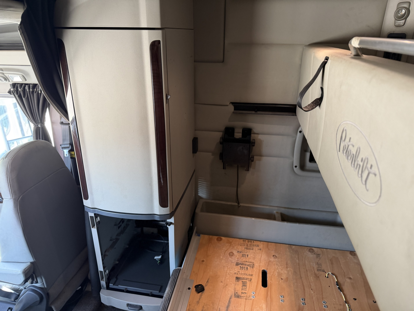 Peterbilt 579 truck interior showing lower cabinet area with a white refrigerator, wooden floor panel, and a sidekick seat. Text visible includes a circular logo on a beige panel and a black switch/box on the wall.