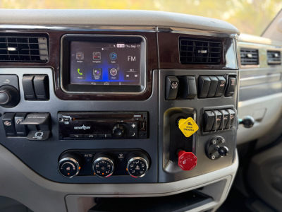 Dashboard of a Peterbilt truck with infotainment screen and control knobs. Visible text includes 'smartnav' on a device, and various control labels. The image shows the center console with climate controls and toggle switches.