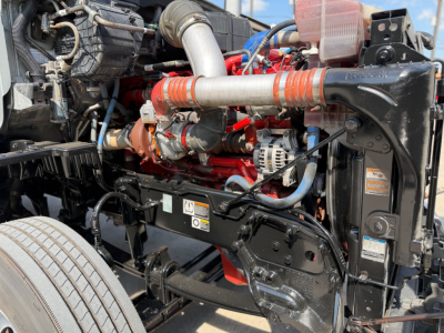Peterbilt 579 engine bay showing red engine block, intake piping, hoses, coolant reservoir, and wiring in front of the cab