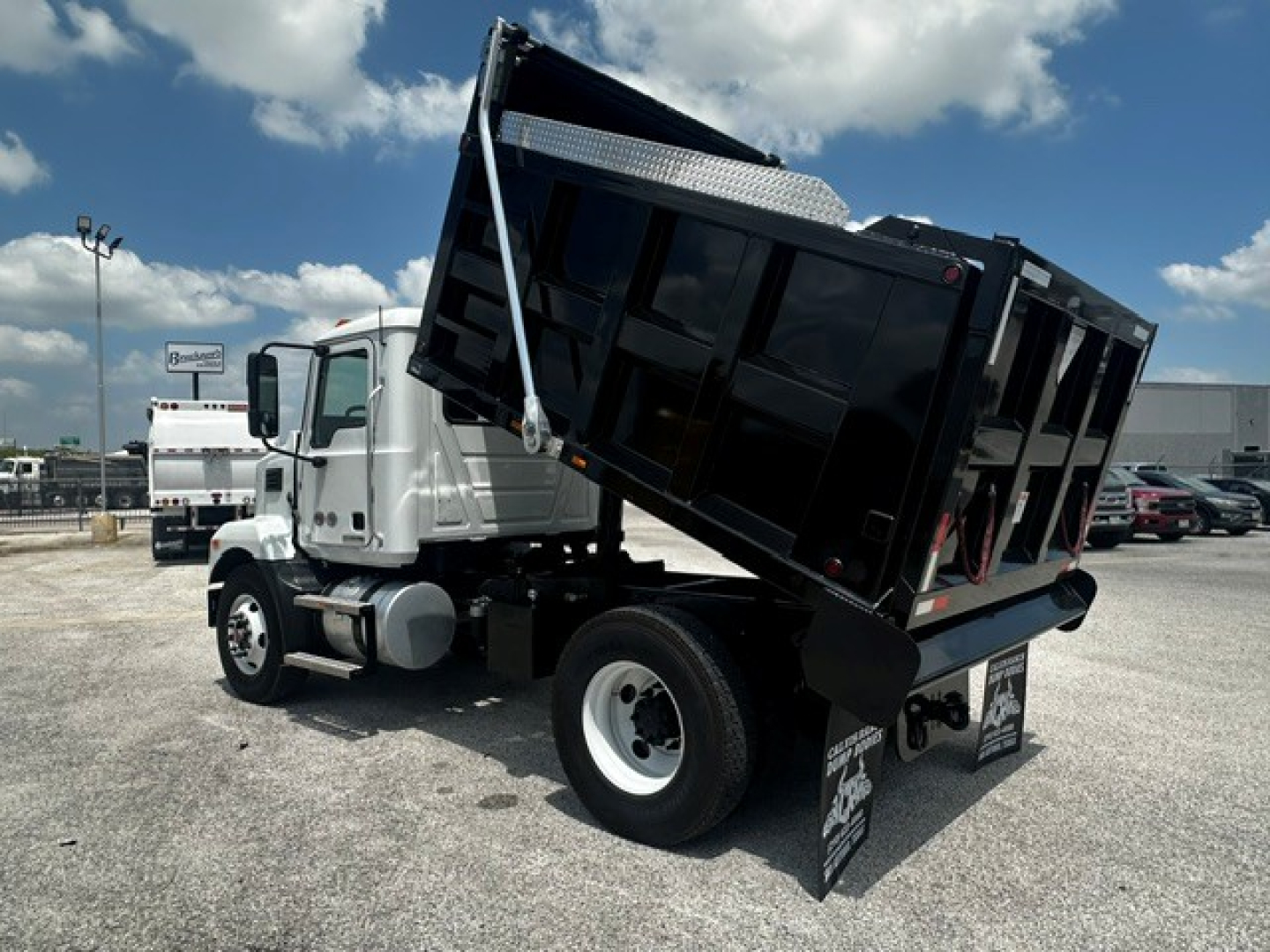 Dump truck in service position showing raised black dump bed on a Mack MD7 chassis with white cab, front wheels visible, gravel lot background.