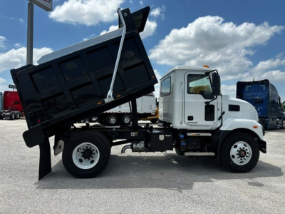 Mack MD7 dump truck in service position with angled dump bed raised, white cab, metal black dump body, side view showing wheel and hydraulic lift.