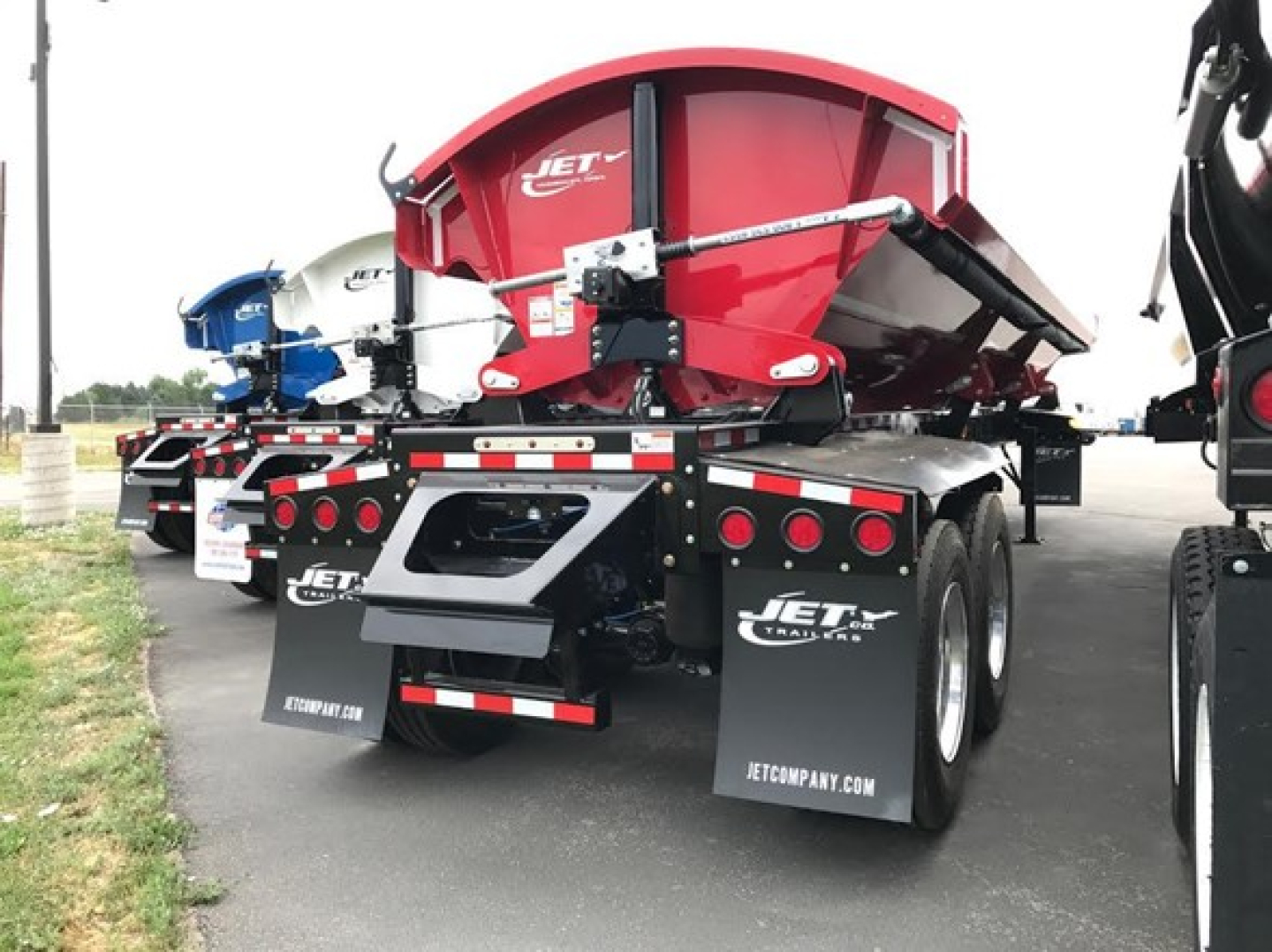 Jet side dump trailer showing red hopper with black frame, dual rear axles, and hanging mud flaps; rear view with reflective tape and hydraulic components visible.