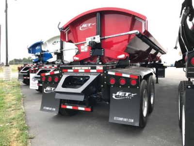 Jet side dump trailer showing red hopper with black frame, dual rear axles, and hanging mud flaps; rear view with reflective tape and hydraulic components visible.