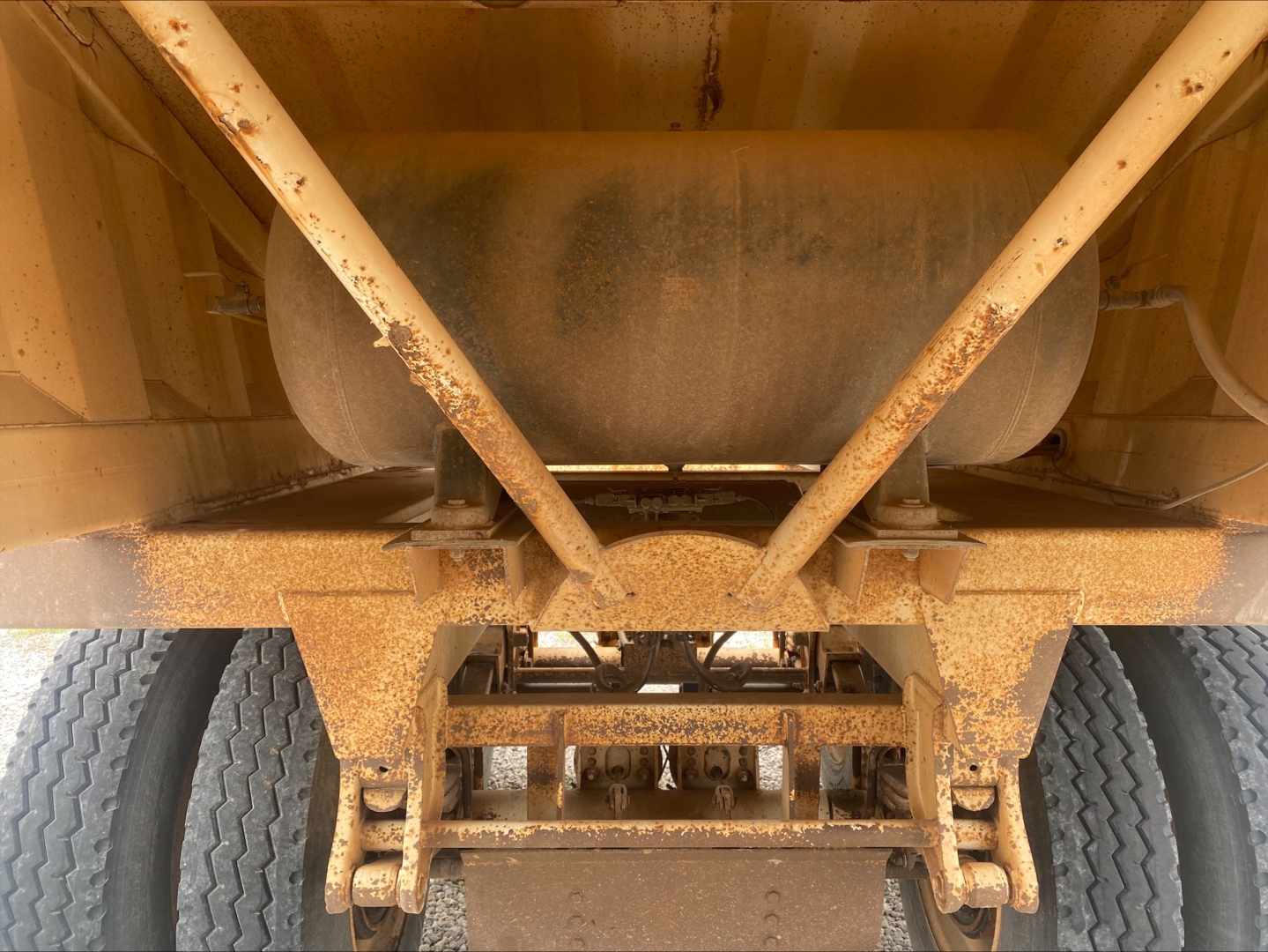 Dump trailer undercarriage with rusted frame and dual rear axles, underside showing suspension and hitch, cylindrical tank and supporting braces.
