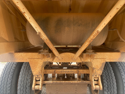 Dump trailer undercarriage with rusted frame and dual rear axles, underside showing suspension and hitch, cylindrical tank and supporting braces.
