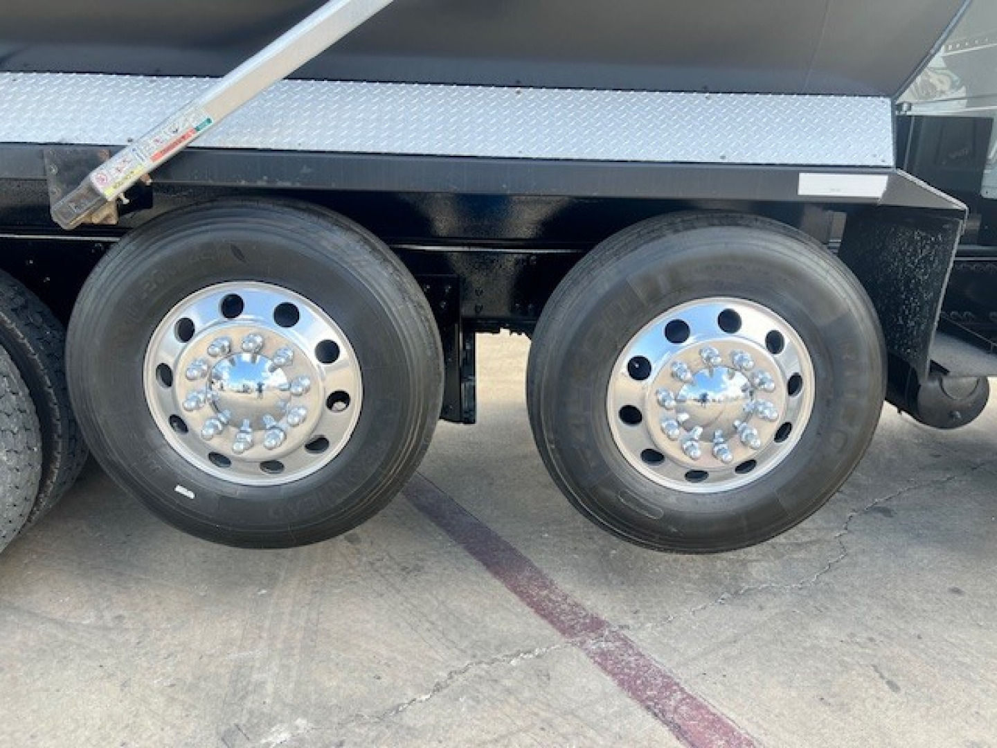 Dual rear axles with polished aluminum wheels on a dump truck; visible are tire treads and a metal step; no legible text other than reflections.
