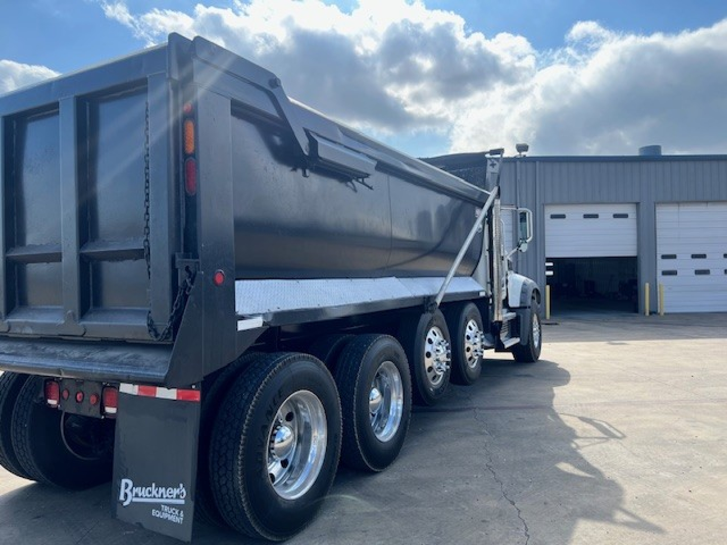 Mack GU713 dump truck rear and side view showing multiple axles, black dump body, and wheel configuration; dealership lot in background.
