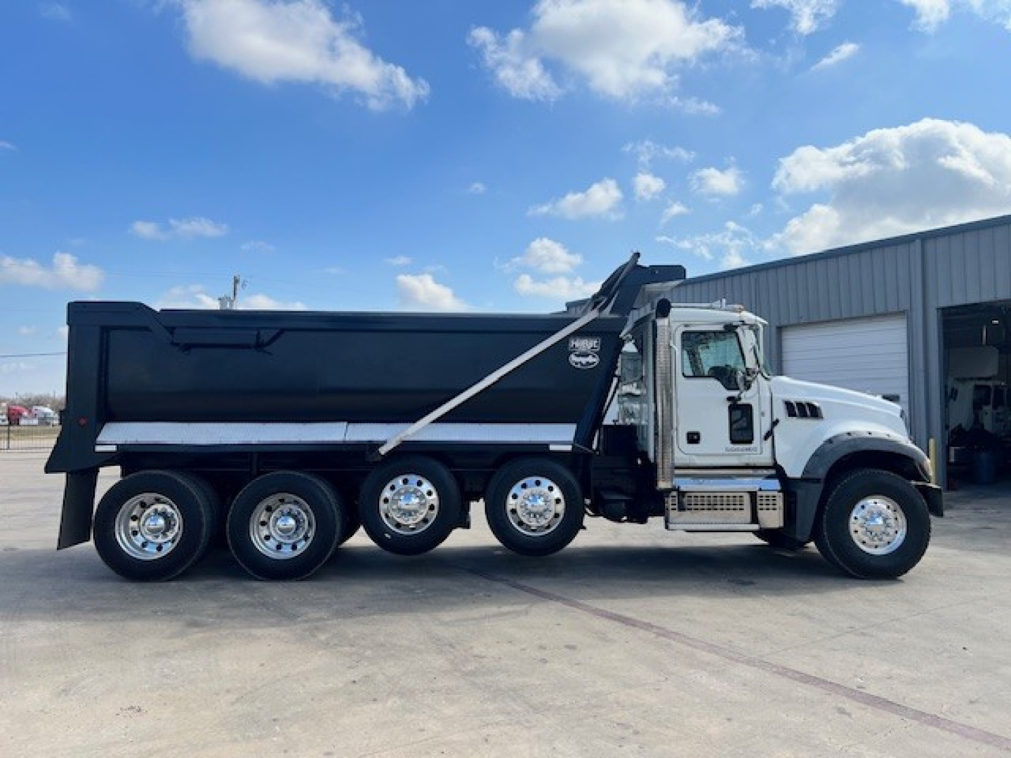 Dump truck side view showing 6-wheel Mack GU713 with black dump body and white cab, under clear sky.