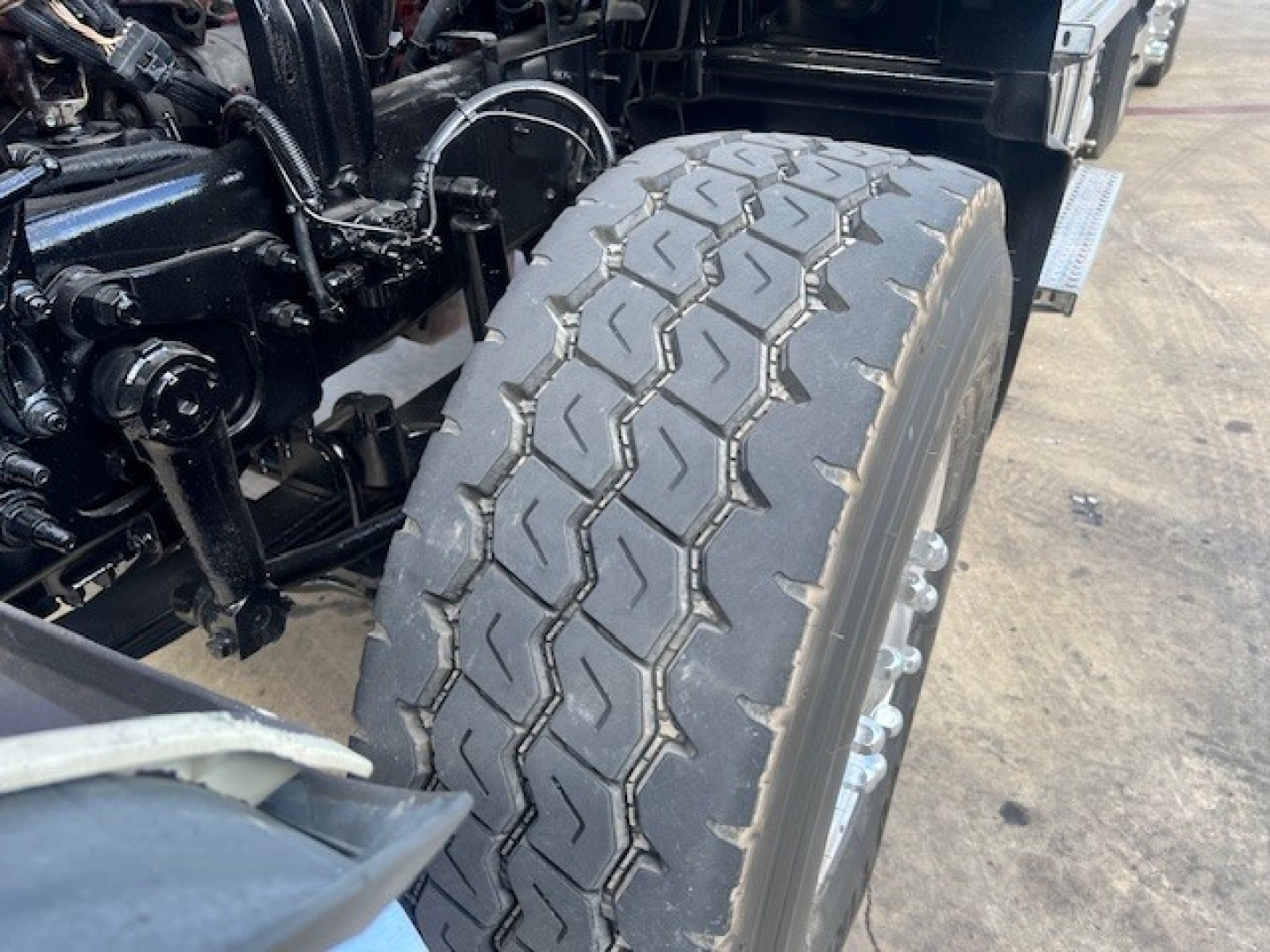 Dump truck front wheel assembly showing large treaded tire and suspension components, chassis, and hydraulic lines visible near wheel well.