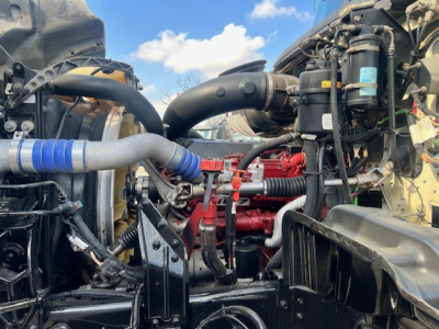 Mack GU713 engine bay view showing red engine block, black hoses, and cooling components; visible labels on filter canisters and wiring harnesses in compact layout.