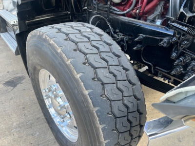 Front left wheel and suspension of a Mack GU713 dump truck showing large treaded tire, chrome wheel, and red engine hoses in the background.