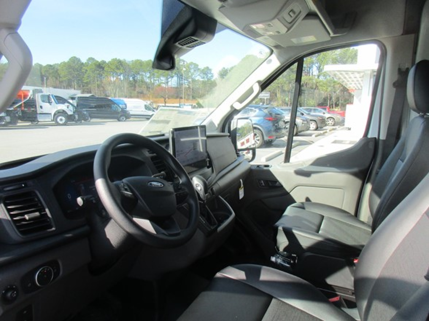 Interior of a 2026 Ford Transit cargo van showing driver seat, steering wheel, and dashboard with center infotainment screen; passenger seat and side window visible.
