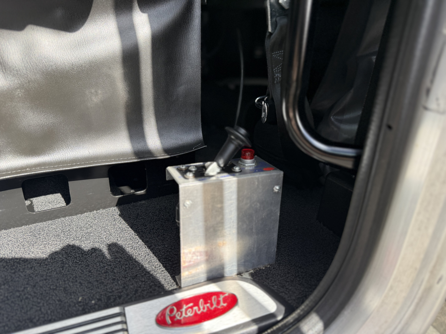 Peterbilt 389 engine bay area showing a metal battery with red terminal cover near the driver seat and floor mat.