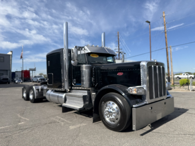 Peterbilt 389 conventional sleeper truck in black with chrome accents, front grille, dual exhaust stacks, and large fuel tanks on a dealership lot.