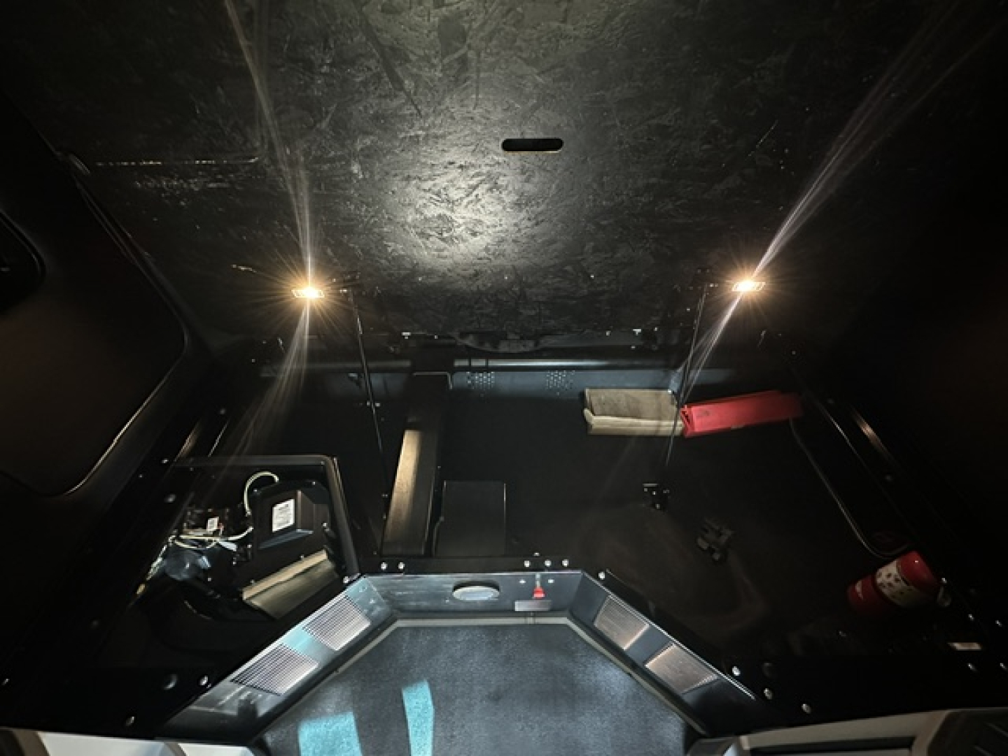 Truck cab interior cargo area/ sleeper of a Kenworth T680 with dashboard not visible; shows black metal walls, small lights, red fire extinguisher and storage area.