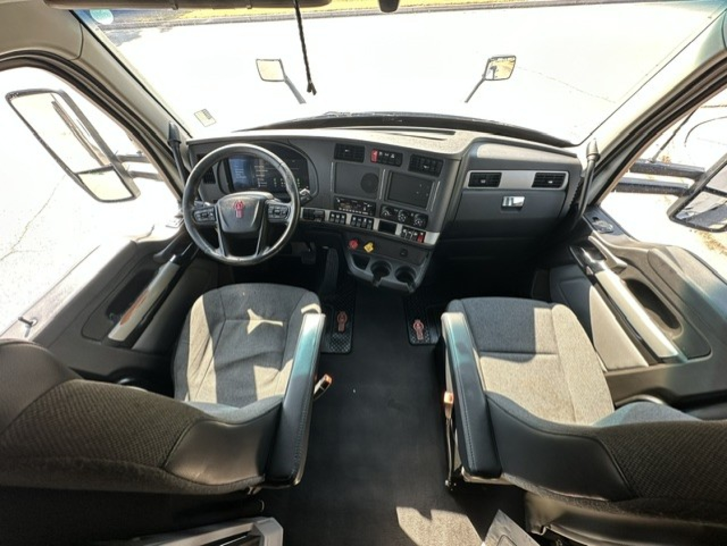 Interior of a 2023 Kenworth T680 cab from the driver perspective, showing steering wheel, dashboard with gauges and controls, two front seats, and center console.