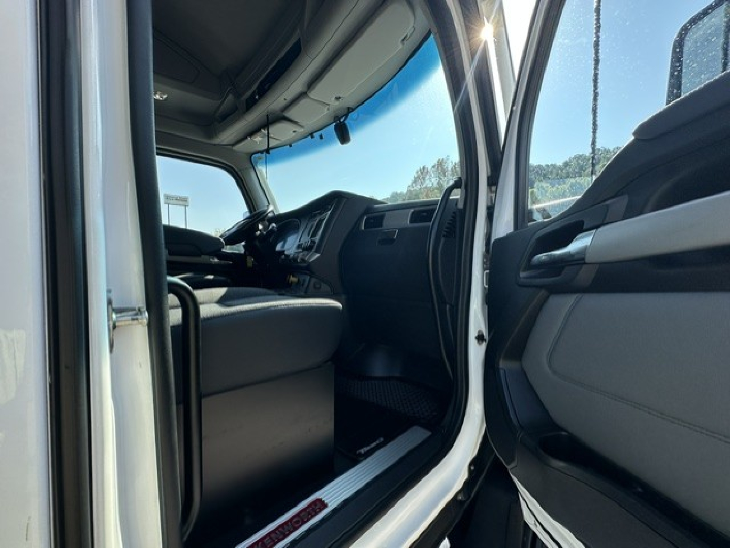 Cab interior of a 2023 Kenworth T680 conventional sleeper truck showing the driver's seat, dashboard, steering wheel, and open door sill