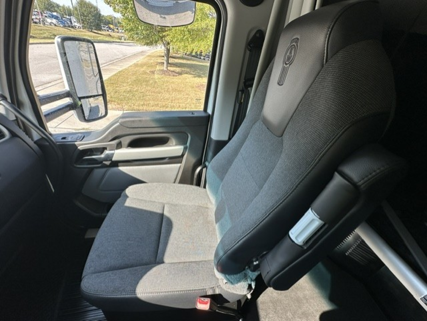 Kenworth T680 truck interior front seat view showing grey fabric seat, armrest, seatbelt buckle, and window/side mirror visible in cab.