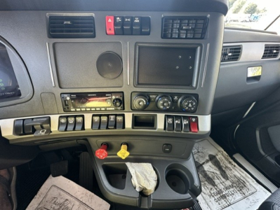 Truck dashboard of a 2023 Kenworth T680 with switches, gauges, and a yellow and red knob; visible text includes no explicit model on panel, but features cup holders and control panels.