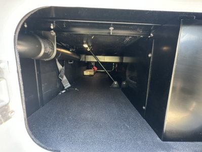 Kenworth T680 truck storage compartment showing under-cab area with wiring, hoses, and a small prop rod; appears to be a storage or access space behind the sleeper.