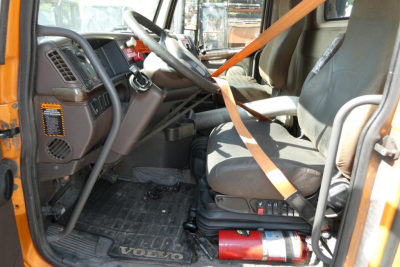  Volvo truck interior with steering wheel, driver's seat, orange seatbelt, and floor mat showing Volvo lettering; dashboard and controls visible.