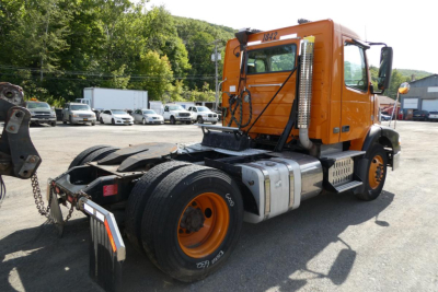 Orange Volvo VNM42T semi truck cab seen from rear-left in a dealership lot, with fifth wheel, fuel tanks, and dual rear wheels.