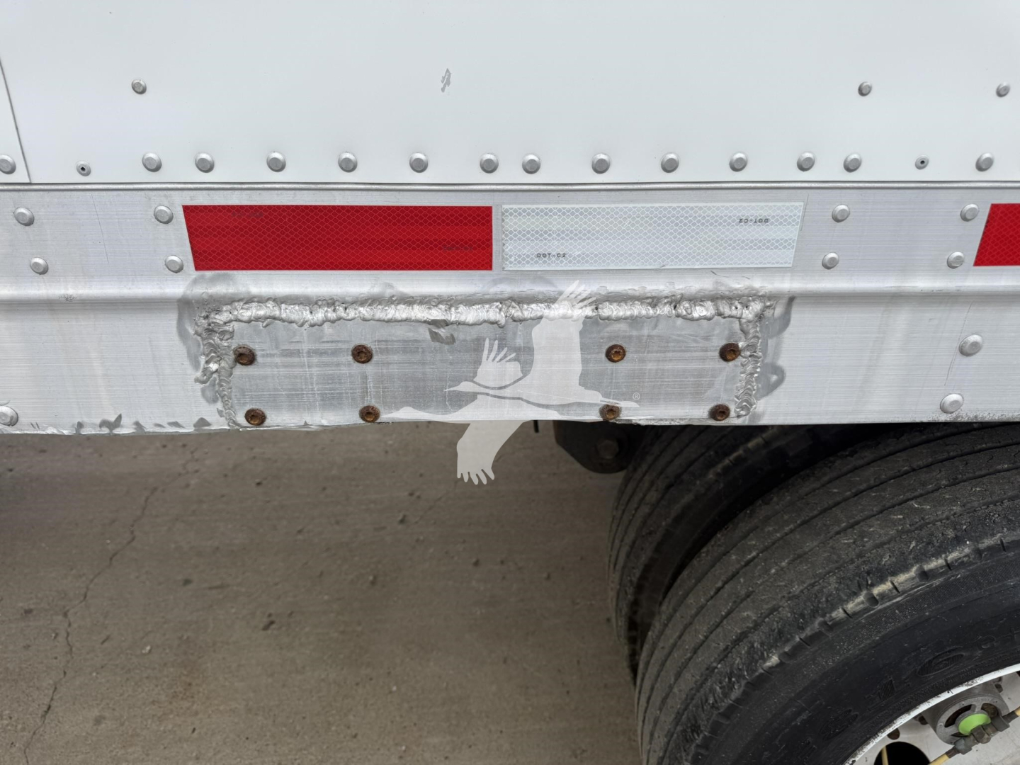 Trailer rear section showing damaged bumper area with riveted aluminum side wall, red and white reflective tape, and dual rear wheels.