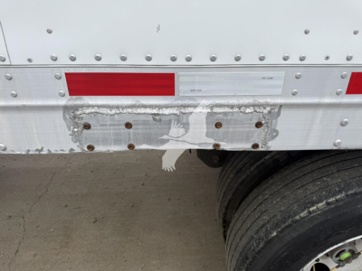 Trailer rear section showing damaged bumper area with riveted aluminum side wall, red and white reflective tape, and dual rear wheels.