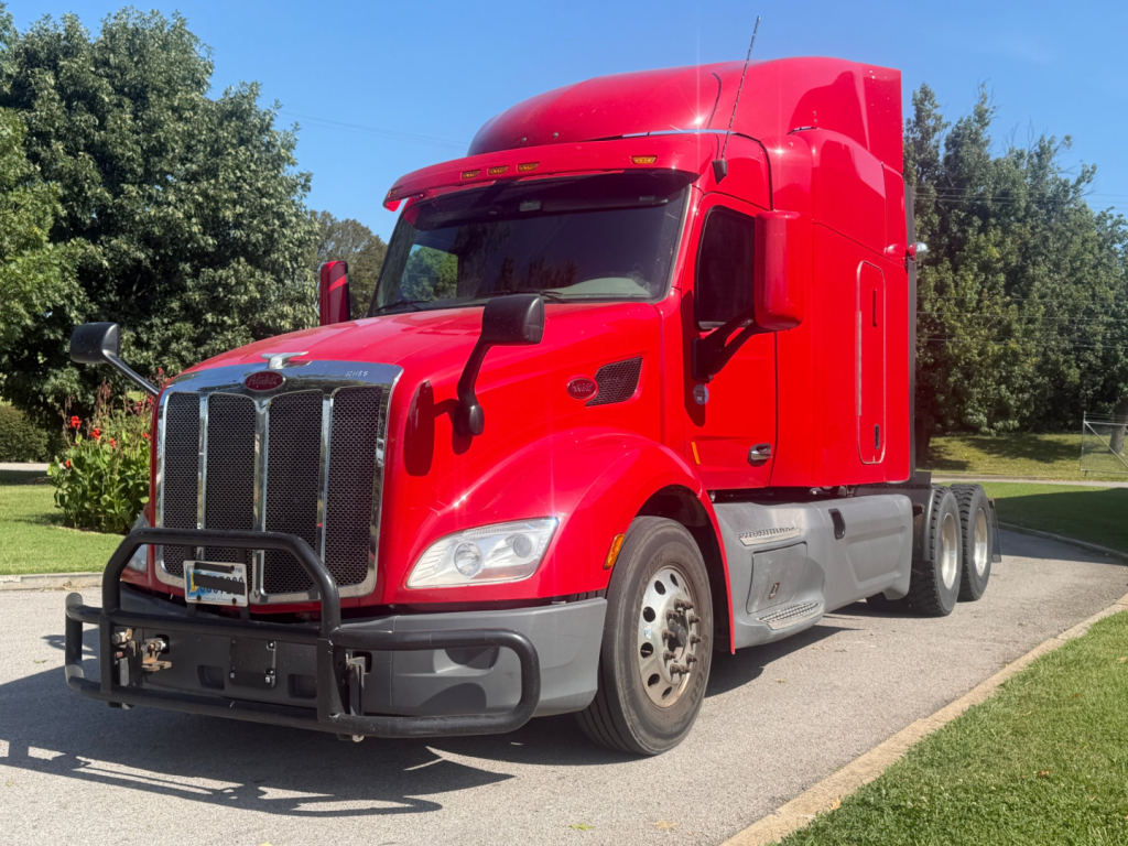 Peterbilt 579 tractor-trailer truck in red, front left view, with grille guard and sleeper cab, daytime outdoor setting.