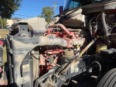 Peterbilt 579 engine bay showing diesel engine with red valve cover and various hoses, radiator, and components visible.
