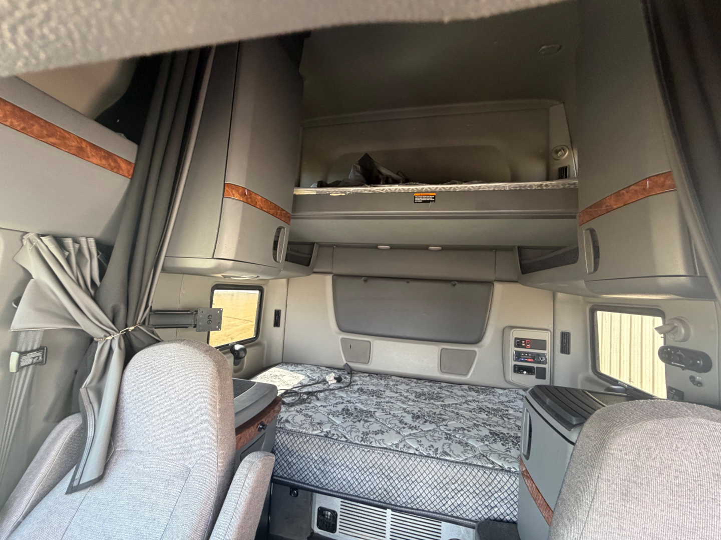 Truck sleeper cab bunk area with overhead storage and ladder steps, interior gray fabric seats, maroon wood trim, and platform bed.
