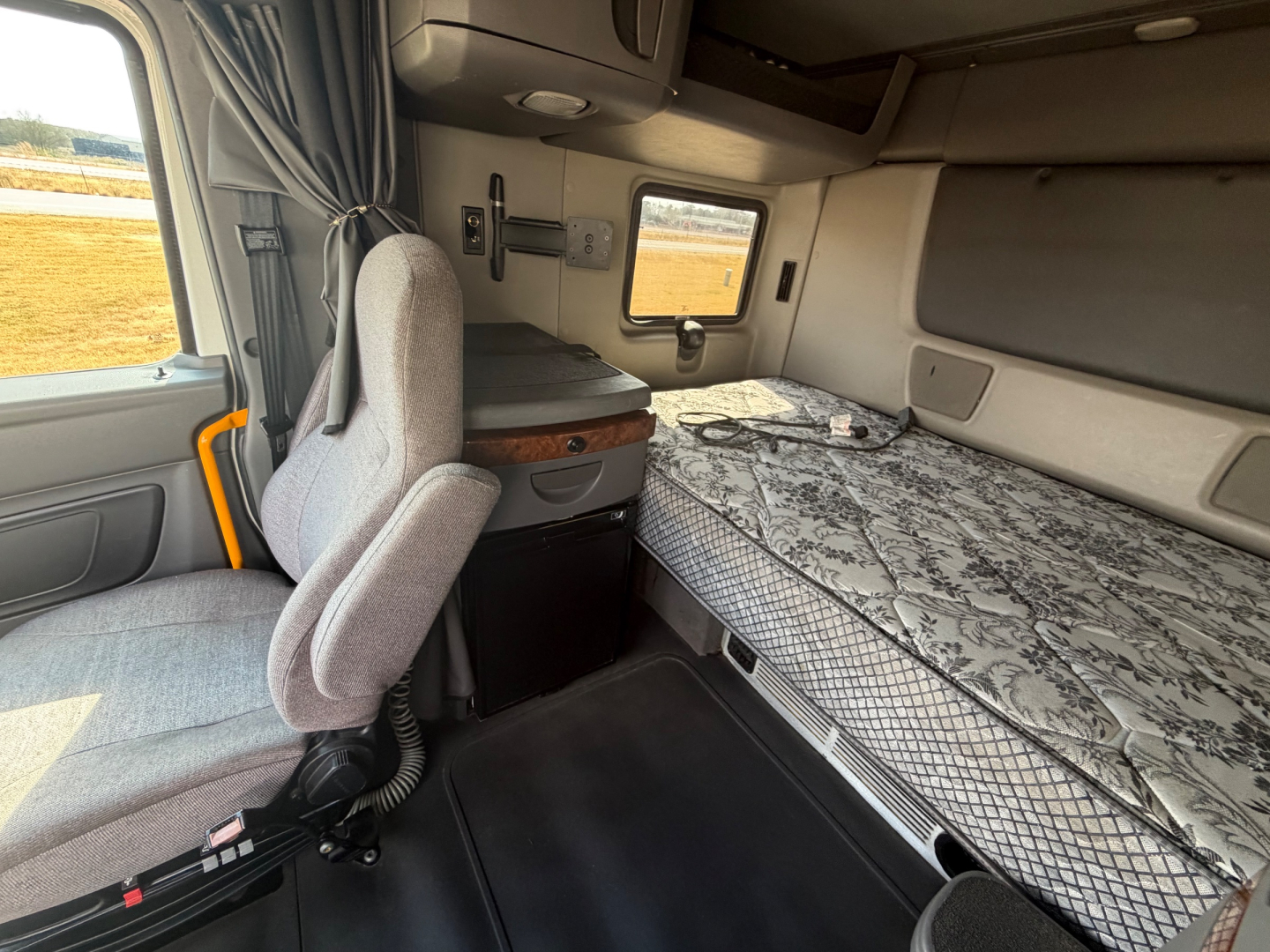 Truck sleeper cabin interior with grey fabric seat, bed with leopard-pattern mattress, small cabinet, window, and overhead storage seen in the sleeper area.