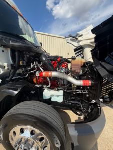 Truck engine bay of a 2021 International LT625 with exposed red and black engine components, coolant reservoir, hoses, and front wheel visible.