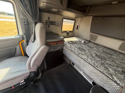 Truck sleeper cabin interior with grey fabric seat, bed with leopard-pattern mattress, small cabinet, window, and overhead storage seen in the sleeper area.