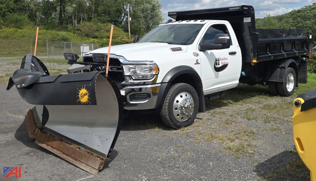 Dump truck with plow attachment, white Ram 5500 single cab, black dump bed, plow blade in foreground, logo on door, daylight outdoor yard.