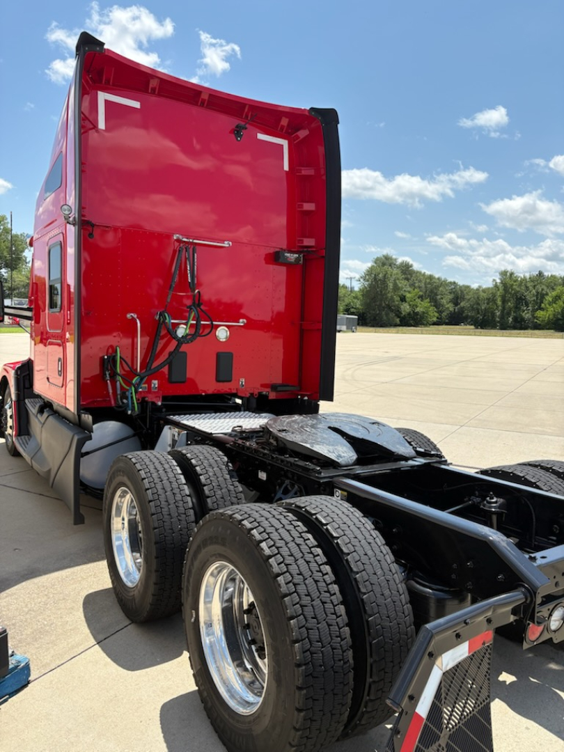 Kenworth T680 tractor rear cab view showing red sleeper with back panel, dual rear axles, and fuel/air lines; clear sky and empty lot in background.