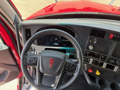 Cab interior of a red Kenworth T680 showing steering wheel with emblem, dashboard gauges, and control panel including switches and vents.