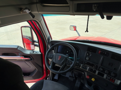Kenworth T680 truck interior view from driver seat showing steering wheel, dashboard, and open door with red exterior.