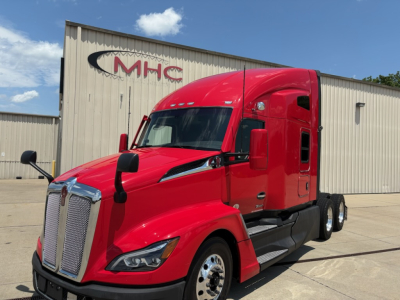 Kenworth T680, red sleeper cab, front left side view, chrome grille, black lower panels, two mirrors, daytime running lights, road grime minimal, background warehouse.
